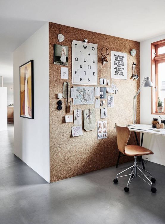 Rincón de despacho con pared de corcho cubierta de notas y fotos, mesa y silla junto a ventana.