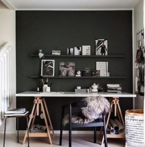 Espacio de trabajo minimalista con escritorio blanco y estanterías oscuras en la pared. Silla negra con cojín de pelaje, luz natural y objetos decorativos.