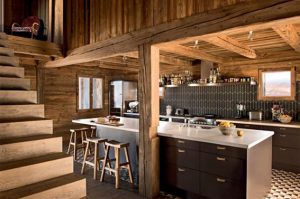 Cocina rústica en madera con isla y barra de cuarzo; taburetes, gabinetes oscuros y encimera blanca, paredes de tronco.