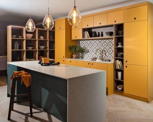 Cocina moderna con muebles amarillos, isla gris y encimera blanca; estanterías abiertas y azulejo decorativo detrás del fregadero.